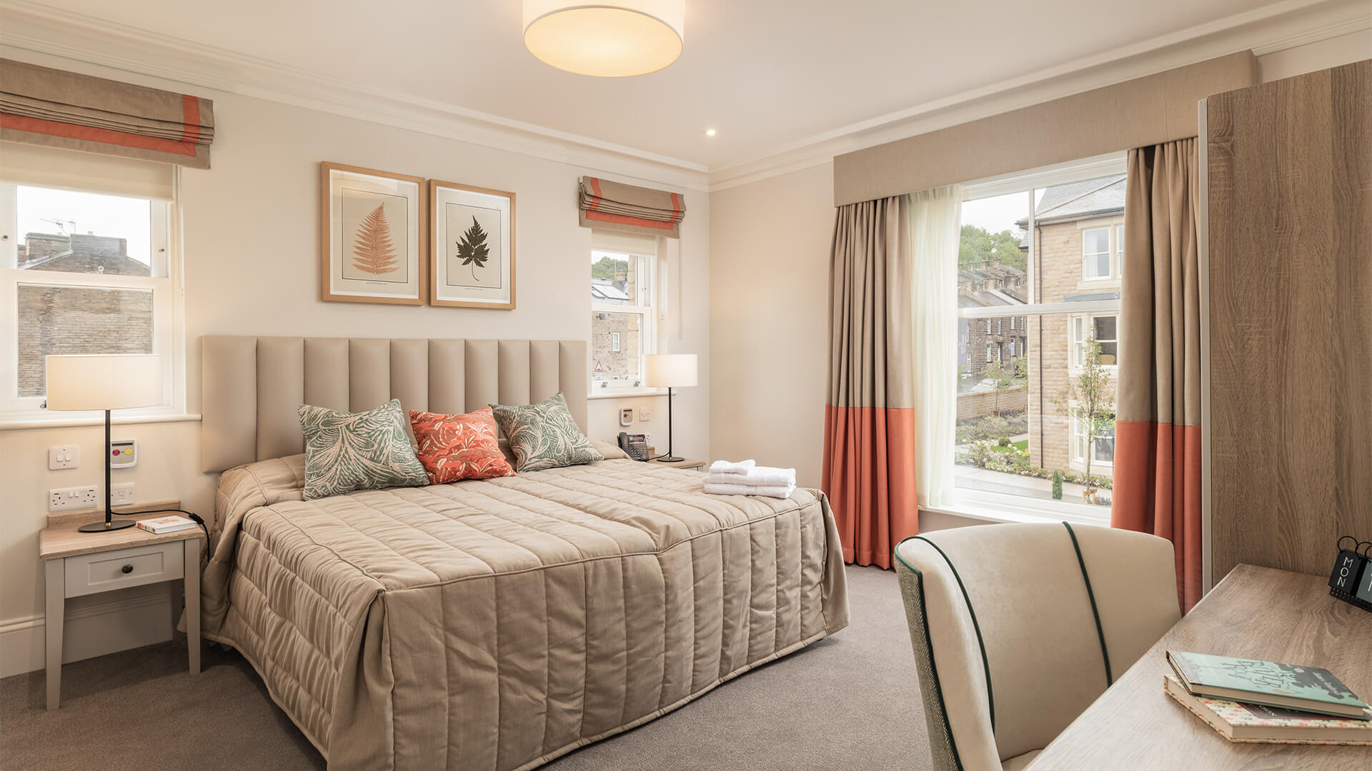 Bedroom with large bed in neutral colours and coral accents. Desk and wardrobes opposite the bed